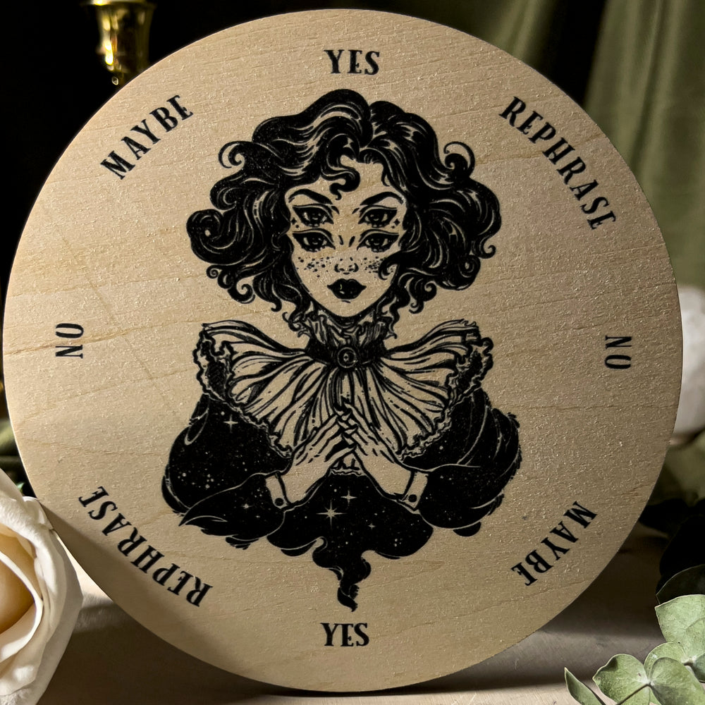 Pendulum board with yes, no, maybe, and rephrase on the edge of the circular board and in the center a witchy woman with four eyes watches.